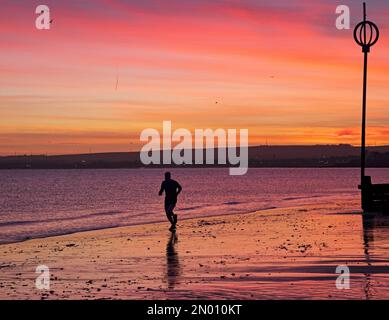 Portobello, Edinburgh, Schottland, Vereinigtes Königreich, 5. Februar 2023. Kühler, farbenfroher Himmel bei Sonnenaufgang über dem Firth of Forth und dem Strand am Meer für die wenigen Menschen, die ihn erleben. Temperatur 1 Grad Celsius mit Eis auf nassen Oberflächen und Autoscheiben. Abbildung: Einsamer Mann, der an der Küste joggt. Kredit: Archwhite/alamy Live News. Stockfoto
