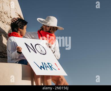 Eine Mutter und ihr asiatischer Sohn sitzen vor einem Bunker und schauen sich an, während sie ein Schild mit der Aufschrift "kein Krieg" halten. Kein Krieg- und Friedenskonzept. Teachin Stockfoto