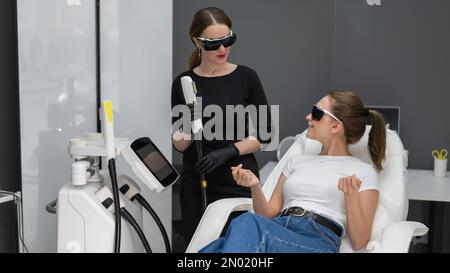 Eine Frau auf einer Couch in einer Brille, die sich einem Fotorejuvenationsverfahren unterzogen hat. Kosmetologe mit einem Patienten. Stockfoto