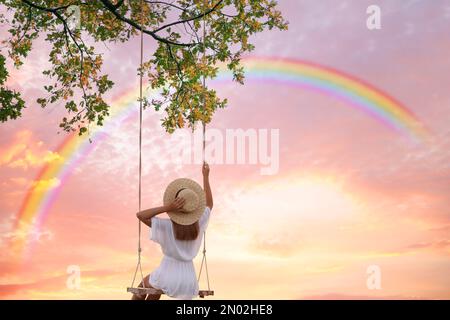 Traumwelt. Junge Frau schaukelt, Regenbogen im Sonnenuntergang im Hintergrund Stockfoto