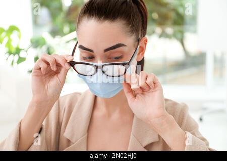 Frau, die neblige Brille abwischte, verursacht durch das Tragen einer medizinischen Maske in Innenräumen, Nahaufnahme Stockfoto