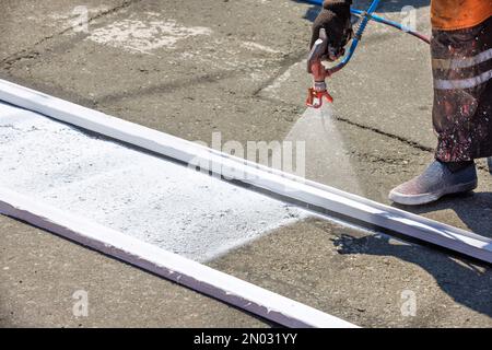 Ein Straßenarbeiter in schmutzigen Overalls trägt an einem Sommertag weiße Straßenmarkierungen mit einer Sprühpistole auf. Speicherplatz kopieren. Stockfoto