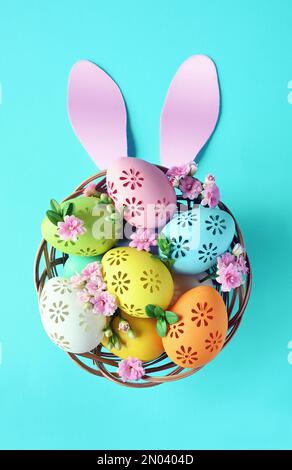 Bunte Ostereier im Korb mit Hasenohren und Blumen auf blauem Hintergrund. Pastellfarbene Ostereier Stockfoto