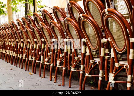 Gestapelte Rattanstühle in einer Reihe in abnehmender Perspektive. Außenterrasse des Restaurants. Steinpflaster. Weicher Hintergrund. Grüner Baum dahinter. Stockfoto