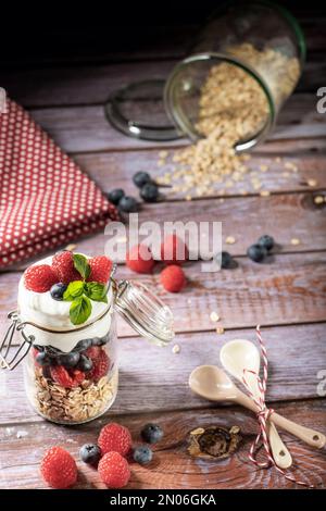 Müsli in einem Glas mit Haferbrei, Himbeeren, Heidelbeeren und Joghurt. Dekoriert auf einem rustikalen Tisch mit Beeren, Löffeln, Handtuch. Stockfoto