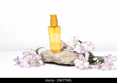 Eine transparente Flasche Öl für Sonnenschutz, Bräunung oder Haarpflege auf einem Podiumsockel. Weißer grauer Hintergrund mit harten Schatten, Blumen und Pflanzen Stockfoto