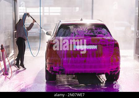 Junge Frau, die ihr Auto mit einem Sprühgerät reinigt. Autowaschanlage mit Selbstbedienung. Orange Auto Stockfoto