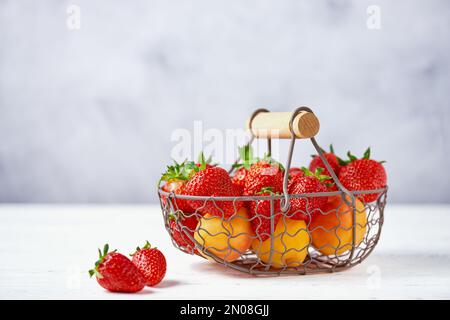 Frische Erdbeeren und Aprikosen in einem Korb aus Metall auf einem weißen Tisch und grauem Hintergrund. Kopierraum, Nahaufnahme. Stockfoto