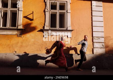 Stylische, fröhliche junge Hipster, bärtiger Mann und blonde Frau halten Händchen und haben Spaß auf den Straßen der alten europäischen Stadt. Ein glückliches verliebtes Paar ist ein Runnin Stockfoto