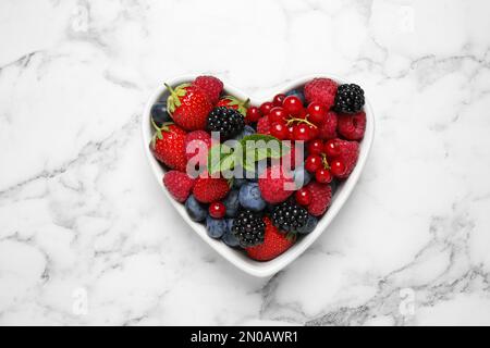 Mischung reifer Beeren in herzförmiger Schüssel auf weißem Marmortisch, Draufsicht Stockfoto