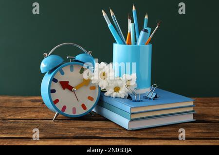 Wecker, Blumen und Schreibwaren auf einem Holztisch in der Nähe der Tafel. Lehrertag Stockfoto