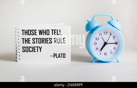 Diejenigen, die Geschichten erzählen, regieren die Gesellschaft, Plato zitiert den alten griechischen Philosophen. Inspirierende Handschrift auf Notizblock auf weißem Hintergrund mit Uhr, Stockfoto