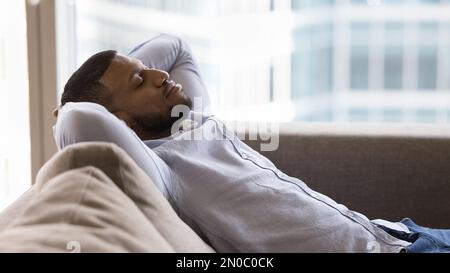 Friedlicher, schläfriger, gutaussehender Schwarzer, der sich auf einer weichen Couch entspannt Stockfoto