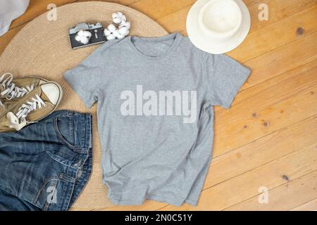 Graues T-Shirt-Mockup mit Baumwollbällen, Jeans und Sneakers, auf Jute-Seilteppich und Holzbrettern im Hintergrund. Stockfoto