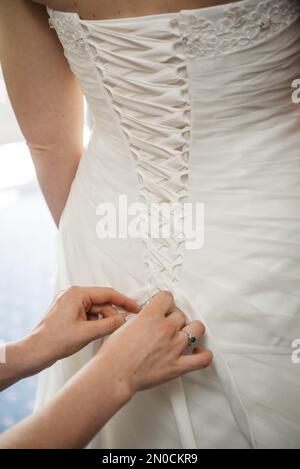 Ein vertikales Bild einer Frau, die einen Schleifenknoten auf der Rückseite des Hochzeitskleides der Braut macht. Stockfoto