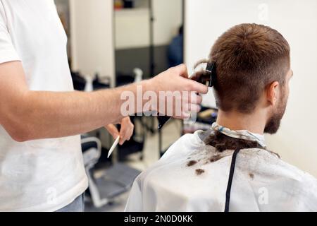 Friseur trimmt Haare mit Haarschneider an einem gutaussehenden bärtigen Mann Stockfoto