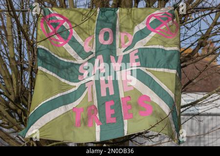Rochford, Großbritannien. Februar 2023. Ein modifizierter Union Jack mit grünem Hintergrund und rosafarbenem „GOD SAVE THE TREES“-Schriftzug mit Logos für Extinction Rebellion, der draußen in Rochford hing. Blattlose Bäume bilden den Rahmen der Szene und betonen die saisonale Dringlichkeit und ökologische Symbolik. Demonstranten von Save Holt Farm Oak Tree protestieren, um eine 100 Jahre alte Eiche zu retten. Der Baum wird voraussichtlich bis April 2023 gefällt. Penelope Barritt/Alamy Live News Stockfoto