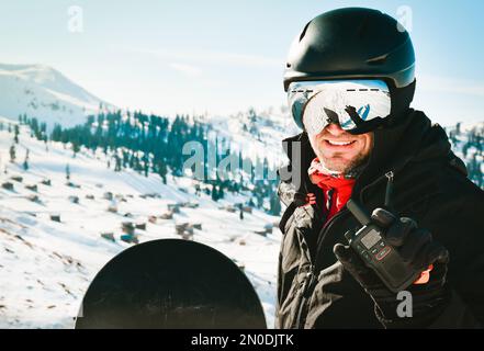 Ein weißer Mann in schwarzer Jacke und Schutzbrille mit Skihelm auf dem Kopf hält ein Walkie-Talkie in der Hand vor dem Hintergrund von weißem Schnee Stockfoto