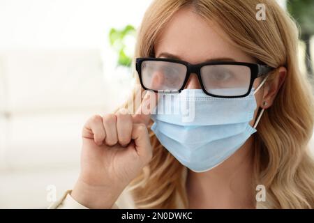 Frau, die neblige Brille abwischte, verursacht durch das Tragen einer medizinischen Maske in Innenräumen, Nahaufnahme Stockfoto