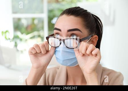 Frau, die neblige Brille abwischte, verursacht durch das Tragen einer medizinischen Maske in Innenräumen, Nahaufnahme Stockfoto