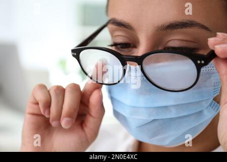 Frau, die neblige Brille abwischte, verursacht durch das Tragen einer medizinischen Maske in Innenräumen, Nahaufnahme Stockfoto