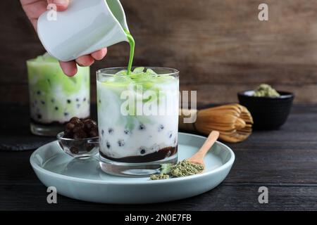 Person gießt grüne Matcha in Glas mit Milchblasentee auf schwarzem Holztisch, Nahaufnahme Stockfoto