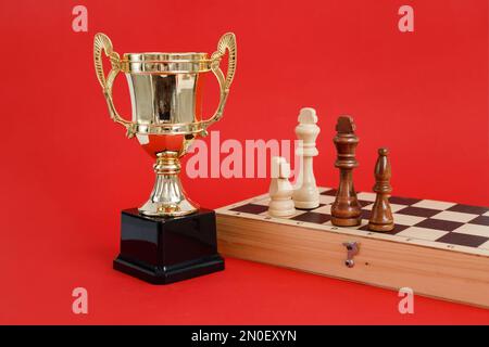 Gewinner des goldenen Pokals und des Schachbretts mit Holzfiguren, die noch leben, auf rotem Hintergrund Stockfoto