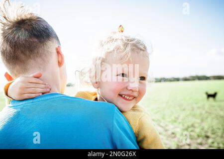 Die kleine Schwester lächelt. Das Mädchen schaut hinter die Schulter ihres Bruders. Ein Teenager trägt ein Kind in Armen über das Spielfeld Stockfoto