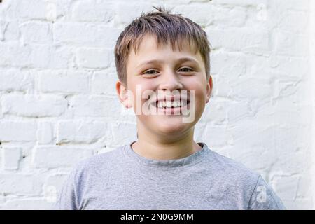 Der Junge lächelt glücklich. Portrait eines unglaublich freundlichen kleinen Jungen. Ein hübscher Teenager in der Nähe einer weißen Ziegelmauer Stockfoto