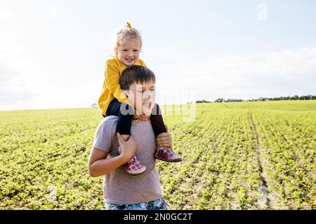 Der Bruder trägt die Schwester auf den Schultern. Kinder laufen draußen auf dem Feld herum. Der ältere Bruder verbringt gern Zeit mit der kleinen Schwester. Beziehungsfamilie ist da Stockfoto