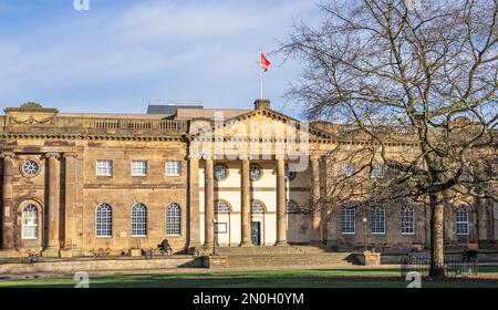 Ein aus Stein gefertigtes Gebäude aus dem 18. Jahrhundert mit Schnitzereien und hohen Säulen im Sonnenschein. Die Leute sitzen auf Bänken und eine Fahne fliegt unter dem Himmel mit Wolken. Stockfoto