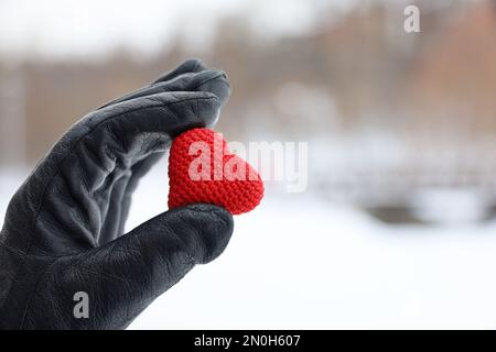 Rotes gestricktes Herz in weiblicher Hand in schwarzem Lederhandschuh auf dem Hintergrund des Schneeparks. Konzept der romantischen Liebe, Valentinstag, Winterwetter