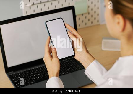 Junge Frau, die ein Smartphone mit weißem Bildschirmmodell hält, während sie am Schreibtisch mit einem Laptop arbeitet Stockfoto