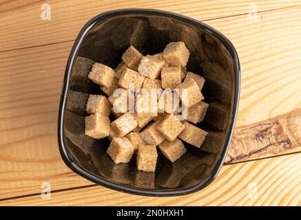 Mehrere Würfel braunen Zuckers auf einem schwarzen Teller, Nahaufnahme, auf einem Holztisch, Draufsicht. Stockfoto