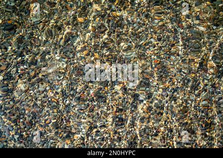 Die Textur des Bodens. Unter dem klaren Wasser sind viele kleine Steine zu sehen. Wasserwellen des Meeres, Sonnenstrahlen auf den Felsen. Stockfoto