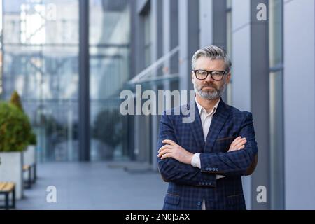 Porträt eines reifen erfolgreichen Chefs, eines grauhaarigen Geschäftsmanns, der mit gekreuzten Armen in die Kamera schaut, eines Mannes außerhalb des Bürogebäudes in Business-Anzug und Brille, eines zufriedenen und fokussierten Bankiers. Stockfoto