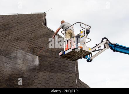 Ein Hausbesitzer, der im Sommer im Freien Moos auf ein Hausdach spritzt und Chemikalien entfernt. Moosentfernungskonzept. Stockfoto