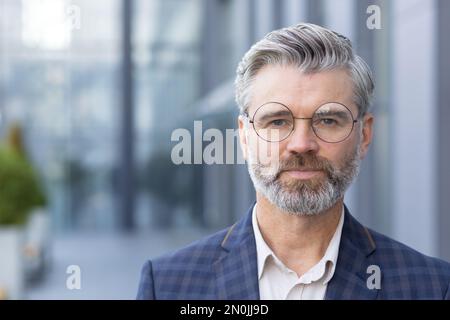Nahaufnahme eines ernsthaften fokussierten Geschäftsmanns, eines erwachsenen Mannes mit Brille und Business-Anzug, der von außerhalb des Bürogebäudes auf die Kamera schaut. Stockfoto