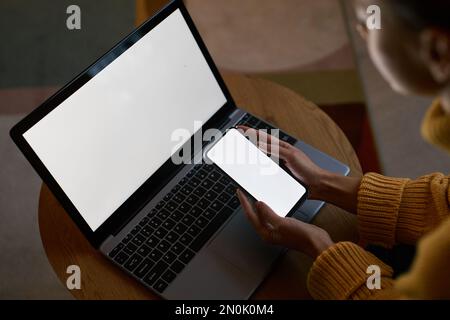 Große Blickwinkel auf eine junge Frau, die Laptop und Telefon mit leeren Bildschirmmodellen verwendet, während sie von zu Hause aus arbeitet Stockfoto