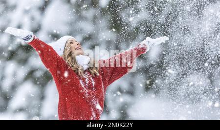 Mit weit geöffneten Armen genießt Frau in rotem Pullover, weißen Handschuhen, Schal und Hut an einem verschneiten Wintertag frische Luft. Stockfoto