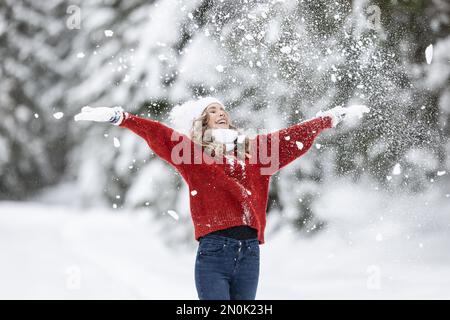 Mit weit geöffneten Armen genießt Frau in rotem Pullover, weißen Handschuhen, Schal und Hut an einem verschneiten Wintertag frische Luft. Stockfoto