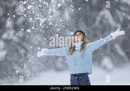 Mit weit geöffneten Armen genießt eine Frau in blauem Pullover, weißen Handschuhen, Schal und Hut an einem verschneiten Wintertag frische Luft. Stockfoto