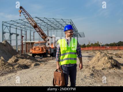 Glücklicher junger Ingenieur mit Helm, der vor der Metallkonstruktion auf der Baustelle steht Stockfoto