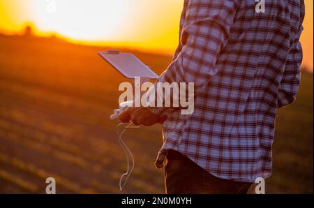 Nahaufnahme der Hände eines Landwirts mit Fernsteuerung zum Fahren der Drohne auf dem Feld am Sommerabend, Innovation in der Agrarwirtschaft Stockfoto