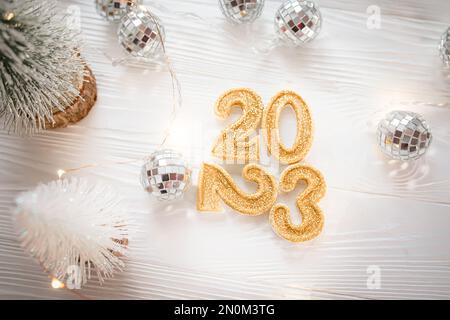 Goldene Zahlen 2023 auf weißem Hintergrund in der Nähe von Discobällen und Weihnachtsbäumen. 2023 kommt im Winter. Heller Hintergrund für das neue Jahr. Stockfoto