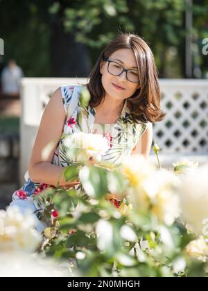 Eine weiße Frau bewundert blühende Rosen im öffentlichen Park. Sommerliche Stimmung. Tropische Pflanzen und Blumen blühen im Garten. Stockfoto