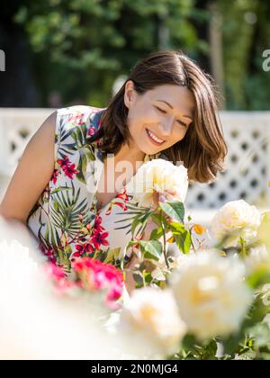 Eine weiße Frau bewundert blühende Rosen im öffentlichen Park. Sommerliche Stimmung. Tropische Pflanzen und Blumen blühen im Garten. Stockfoto