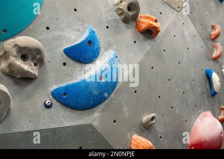 Nahaufnahme von Kletterwänden und Griffen an grauen Kletterwänden im Innenbereich. Farben: Blau, Rot, Orange und Grau. Stockfoto