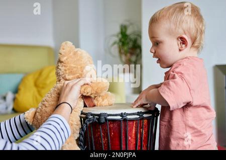 Ein Kleinkind und ein Teddybär spielen zu Hause zusammen Bongo-Trommeln Stockfoto
