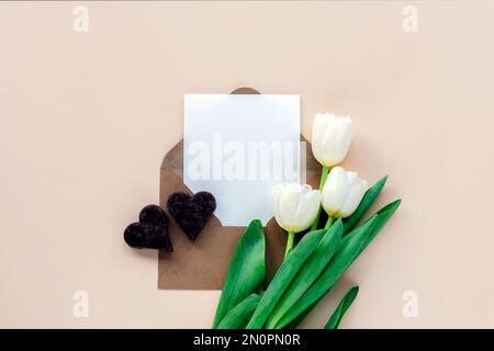 Weiße Tulpen, Umschlag mit leerem Blatt und Schokoladenherzen auf neutralem beigefarbenem Hintergrund. Valentinstag, Muttertag-Feiertagskonzept. Draufsicht, Fla Stockfoto